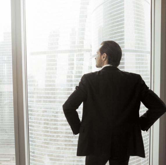 A businessperson in a suit stands with their hands on their hips, looking out of a large window at a cityscape with tall glass buildings. Their posture conveys confidence and forward-thinking, aligning with the concept of a positive mindset in entrepreneurship.